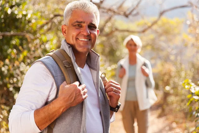 Smiling senior man hiking with backpack in the foreground, woman in background on a sunny trail.