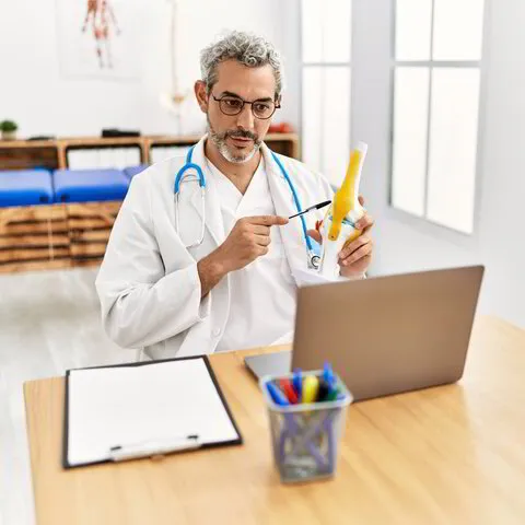 Middle age grey-haired man doctor having video call holding model of knee joint