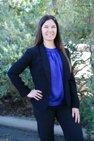 Woman with dark brown hair wearing black business suit and purple blouse standing in front of desert landscape greenery.