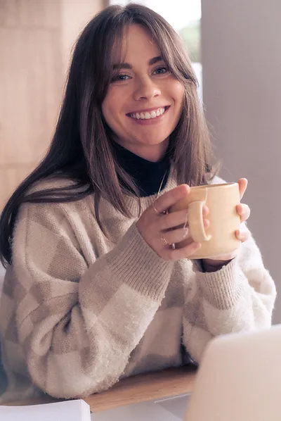 Naomi Murray, RHNin a sweater smiling at the camera, holding a mug, with a laptop in front of her.