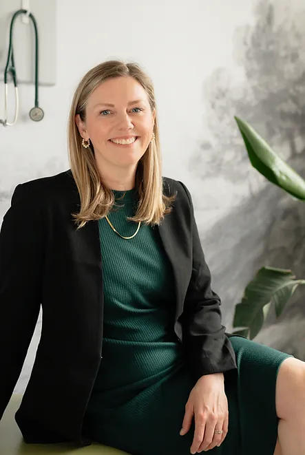 Dr. Akehurst smiling in a clinic office, wearing a black blazer and green dress, with stethoscope and plant in background.