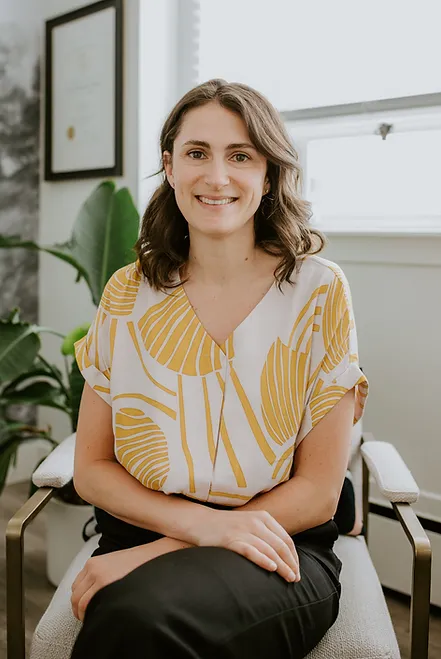 Dr. Brett Simpson, ND, seated in an office, wearing a yellow leaf-patterned blouse and black trousers.