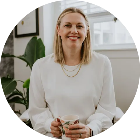 A smiling woman in a white blouse holding a coffee mug, sitting in a well-lit room with plants.