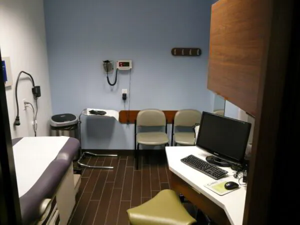 A modern doctor's office with an examination table, computer desk, chairs, and medical equipment on the wall.