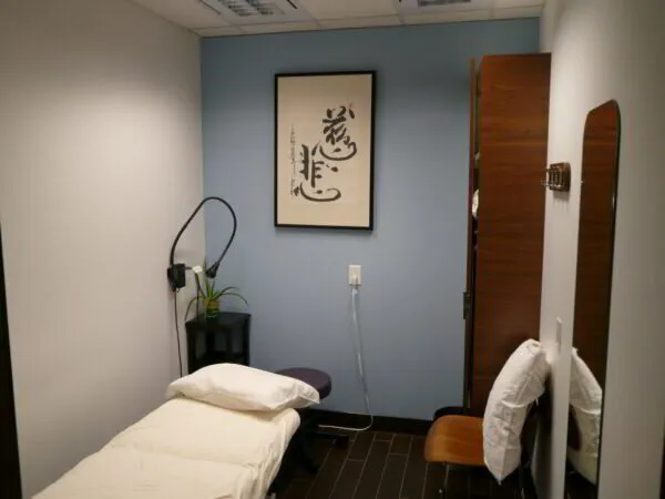 A clean medical examination room with an examination bed, otoscope, chair, and a framed calligraphy artwork on the wall.