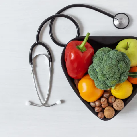 A stethoscope and a heart-shaped bowl filled with fresh vegetables and fruits on a white surface, symbolizing healthy eating.