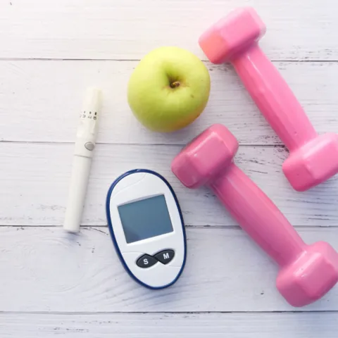 An apple, pink dumbbells, a glucose meter, and an inhaler on a wooden surface, symbolizing health management.