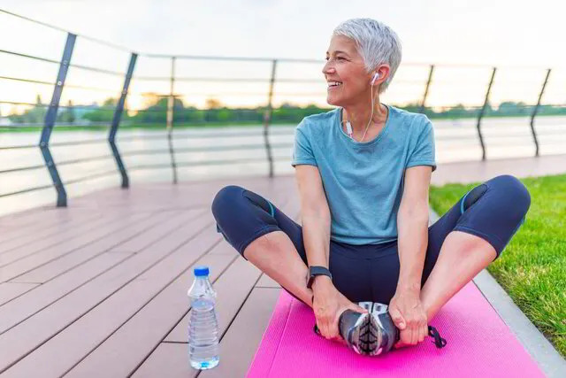 A smiling woman with grey hair stretching on a pink yoga mat outdoors near a river at dusk, with water bottle beside her.