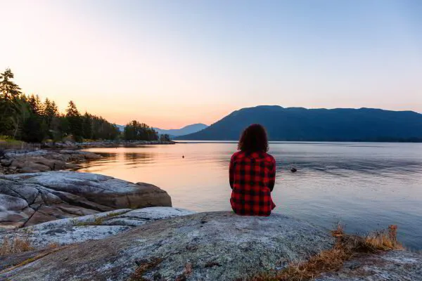 Person in red plaid shirt sitting on a rock, watching a serene lake at sunrise with mountains in the background.