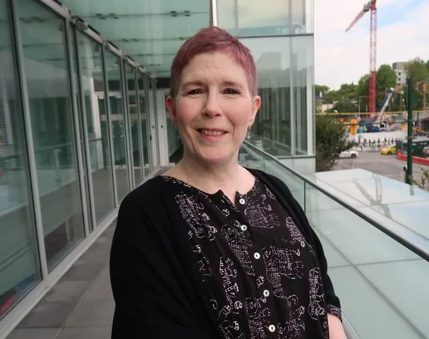 Woman with short red hair smiling on a glass balcony, urban backdrop with trees and construction crane.