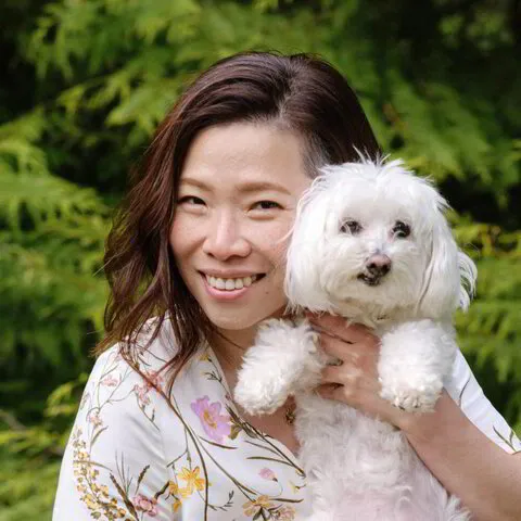 Woman smiling and holding a white fluffy dog outdoors with greenery in the background.