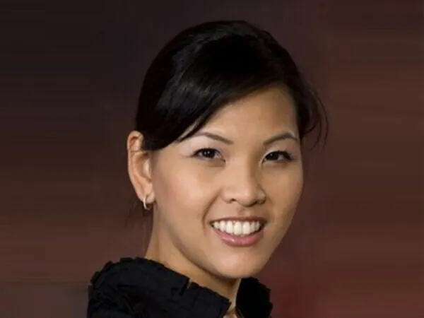 Smiling Asian woman with black hair, wearing a dark blouse, against a blurred brown background.