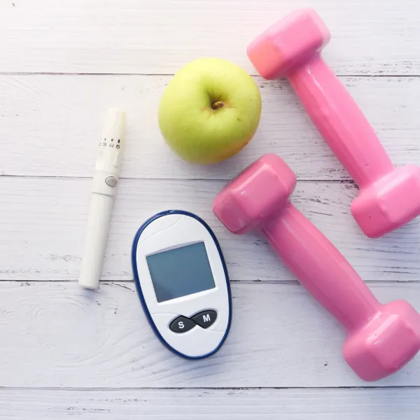 A green apple, pink dumbbells, a glucose meter, and an insulin pen on a wooden surface, symbolizing health management.
