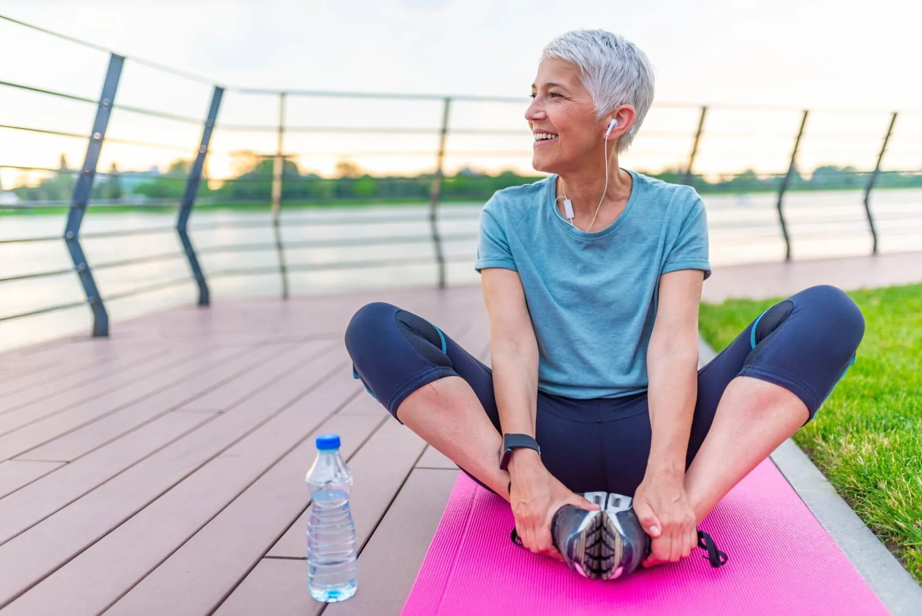 Smiling woman stretching on a yoga mat outdoors with a water bottle beside her.