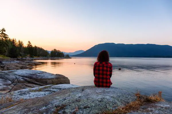 Person in red plaid shirt sitting on a rock by a calm lake at sunset with mountains in the background.