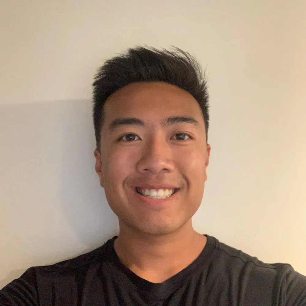 Smiling man with short hair posing for a selfie in a room with a plain wall.