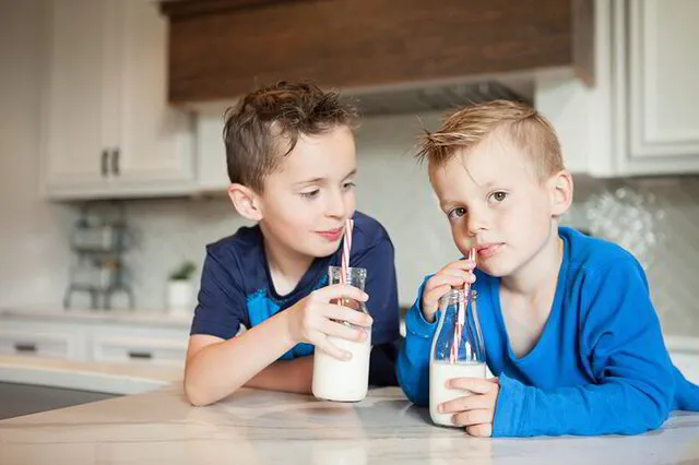 Two young boys drinking milk from glass bottles in a modern kitchen setting.