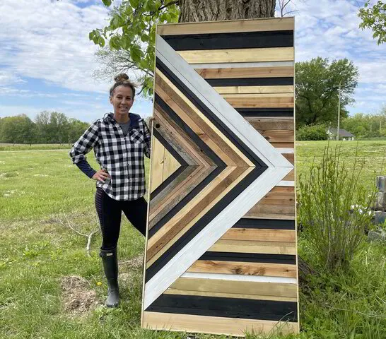 Woman in plaid shirt standing next to a handmade wooden chevron panel outdoors.