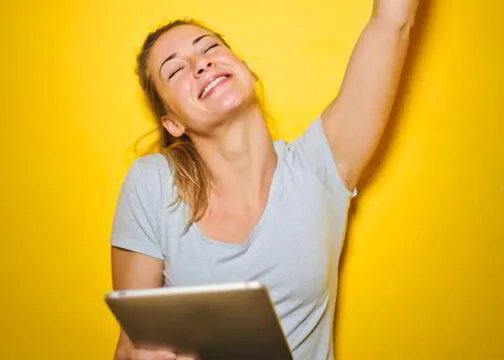 Woman laughing with a tablet in hand, raising her arm in triumph against a yellow background.