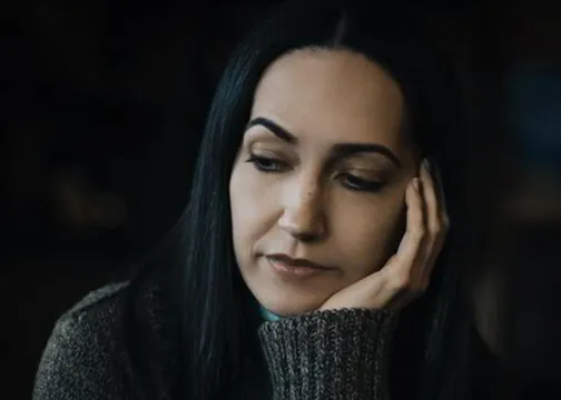 Woman in a gray sweater resting her head on her hand, looking thoughtful.