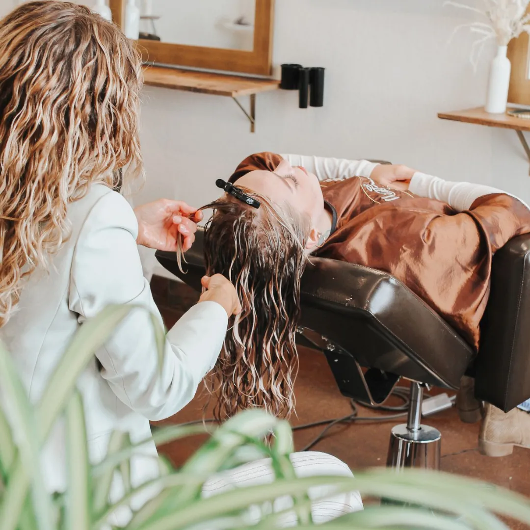 A stylist from behind, with blonde highlighted curls, is carefully trimming the wet hair of a client who is lying back in a salon chair with a shampoo cape around them. The stylist's focus is on achieving the perfect cut to enhance the natural curl pattern, indicative of the specialized services provided at Mid Town Curls salon.