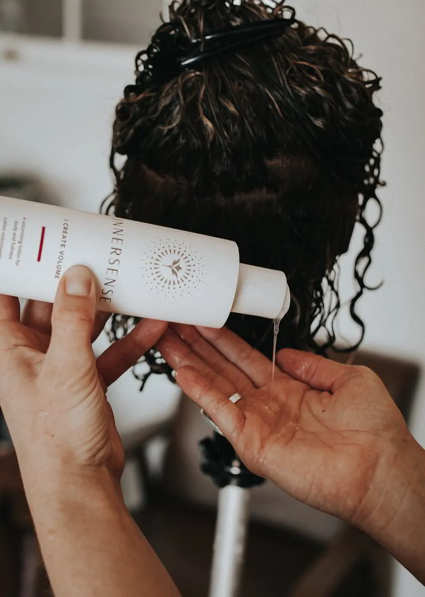 A stylist's hands are pouring a clear, viscous liquid from a white Innersense 'I Create Volume' bottle into their palm, against the backdrop of a client's damp, dark curly hair, ready for the styling product to be applied.
