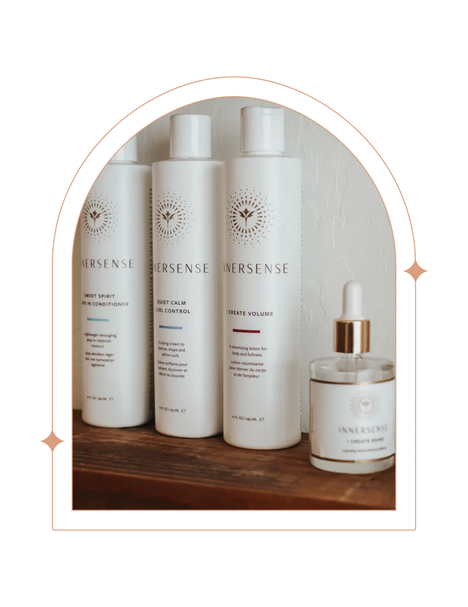 A curated selection of Innersense organic hair care products is neatly displayed on a wooden shelf against a white textured wall. From left to right, there's a 'Sweet Spirit Leave In Conditioner,' 'Quiet Calm Curl Control,' and 'I Create Volume,' all in white bottles with minimalist design, along with a smaller container of 'Harmonic Healing Oil' with a dropper.