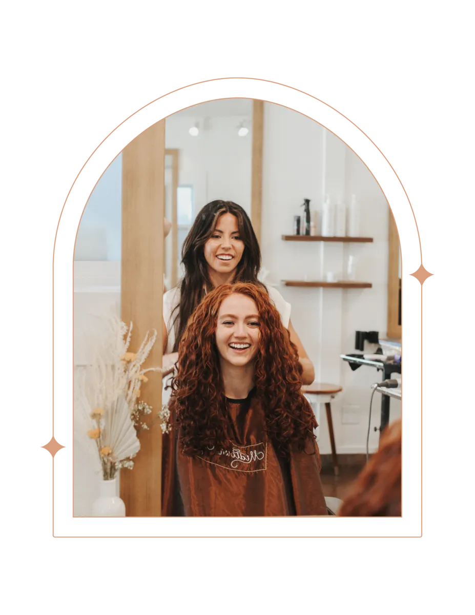 A cheerful stylist stands behind a seated client with vibrant, voluminous, shoulder-length red curly hair at Mid Town Curls salon. The stylist, with dark hair and a bright smile, appears to have just finished styling the client's hair. The client, wearing a salon cape and with a joyous expression, shows off her bouncy curls. They are surrounded by a bright and airy salon interior with stylish decor and a shelf of hair care products in the background.