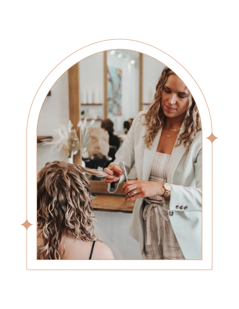 In the warm ambiance of Mid Town Curls salon, a focused stylist with curly hair, dressed in a white blazer, is meticulously trimming the ends of a client's highlighted, shoulder-length curly hair. The client, seen from the back, is looking down, possibly admiring the salon's chic décor in the reflection of a large mirror that reveals a glimpse of the salon's elegant and serene styling area.