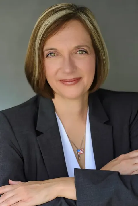 A woman with her arms crossed looking at the camera wearing an American flag necklace and gray suit