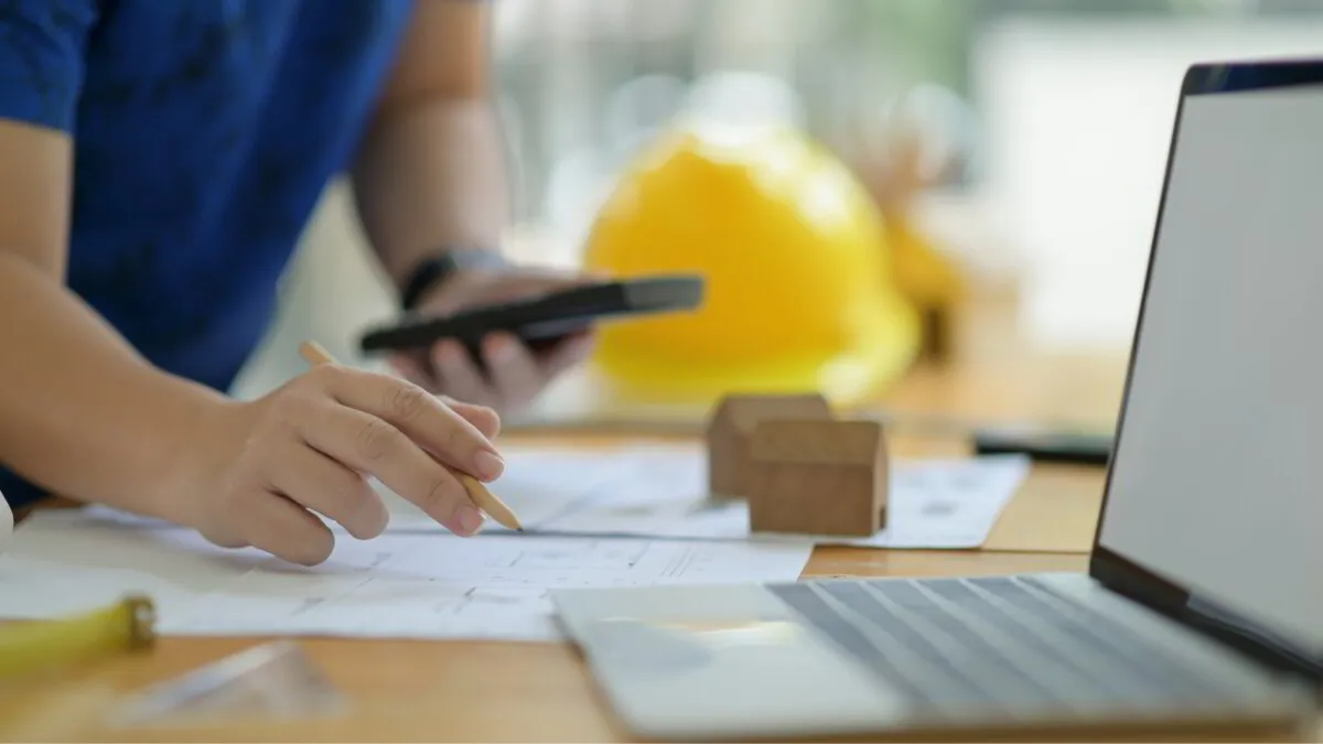 Engineer hands holding pencil and calculator looking at plans on desk with laptop and hard hat