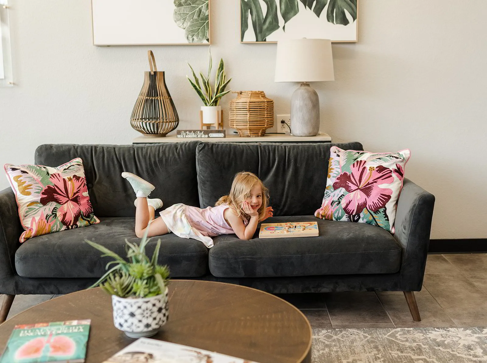 A young girl enjoys her time, playfully reading a puzzle book on a comfy gray sofa, surrounded by vibrant decor and houseplants, feeling at ease