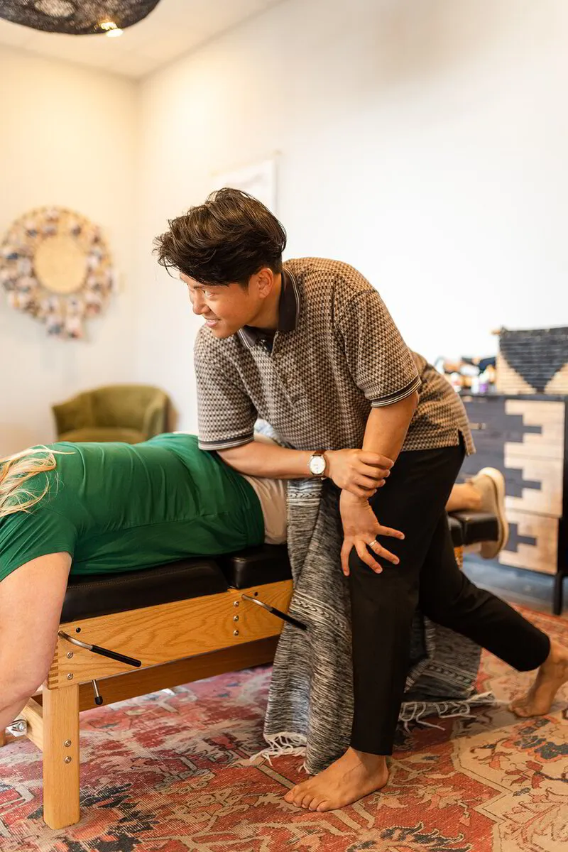 Dr. Binh, dressed in a checkered shirt, carefully assesses his patient's alignment during a hands-on chiropractic session in a cozy clinic