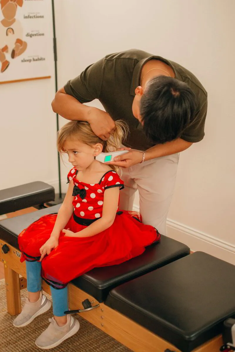A chiropractor attentively uses a handheld device on a young girl in a polka-dot dress