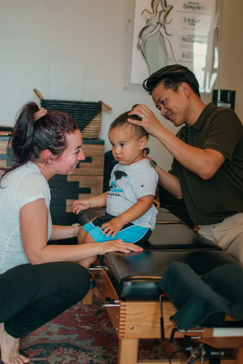 Dr. Binh gently examines a toddler's posture with careful hands, while a woman, likely the mother, watches with a tender smile, creating a picture of trust and care