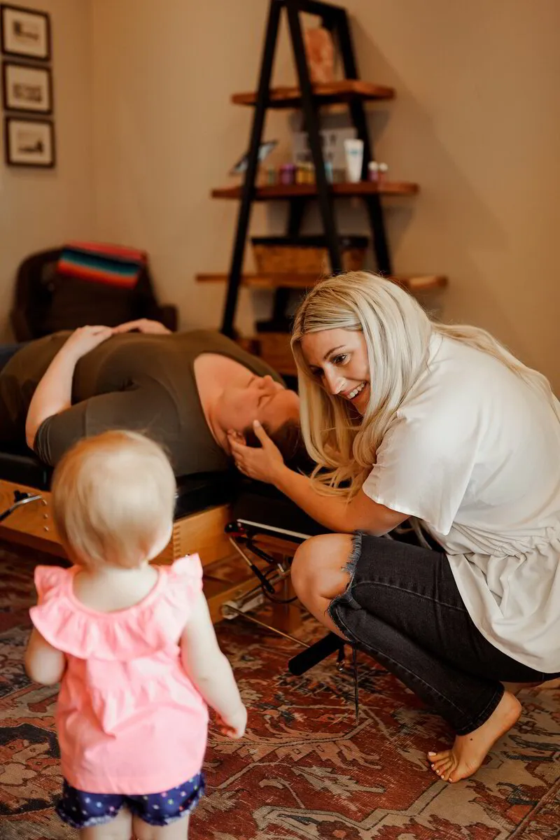  A smiling chiropractor kneels down to interact with a toddler while treating a patient who is lying down on a chiropractic table in a warm, welcoming room