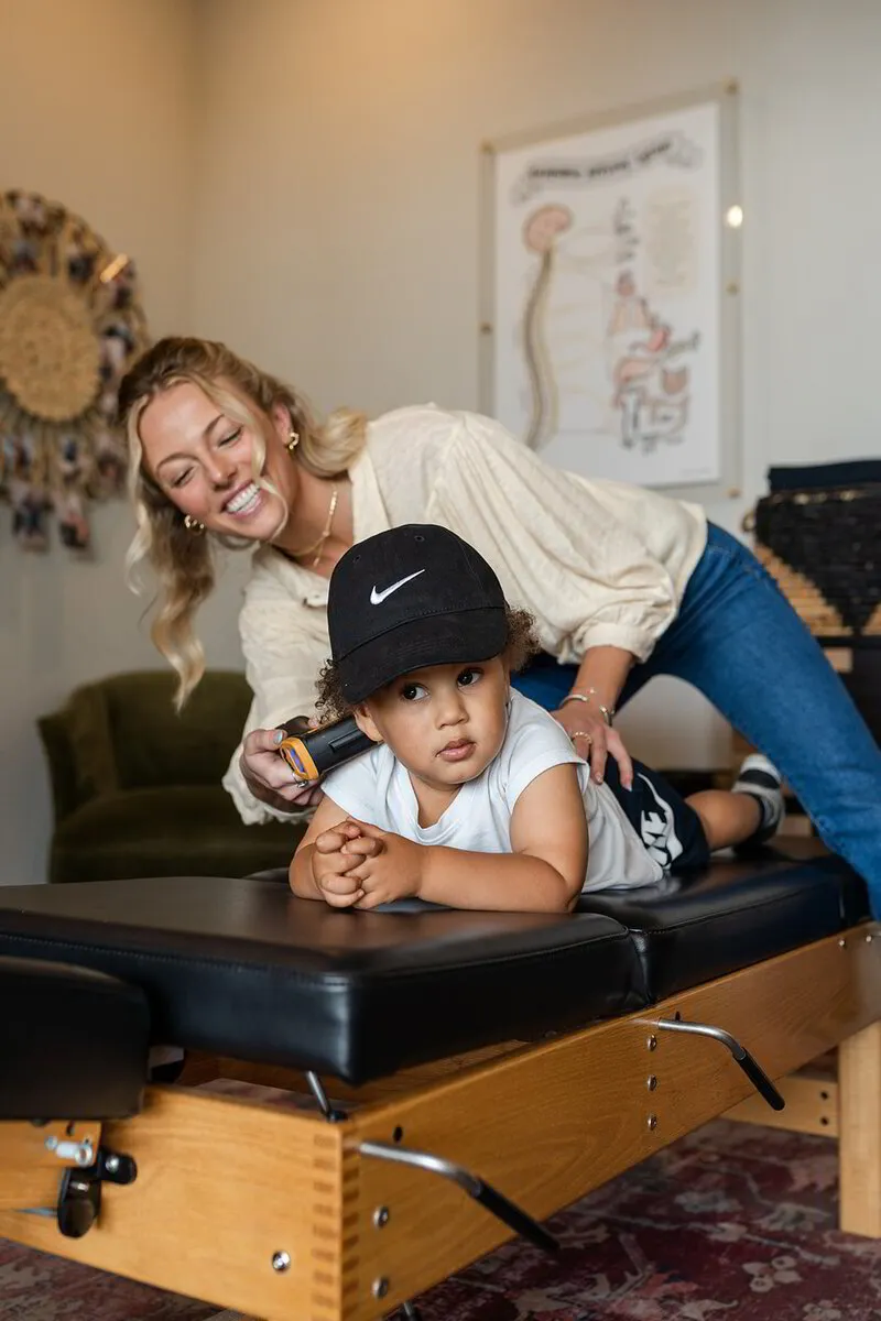  Dr. ALEXIS engages with a young child on a chiropractic table, her bright smile reflecting a warm, reassuring atmosphere