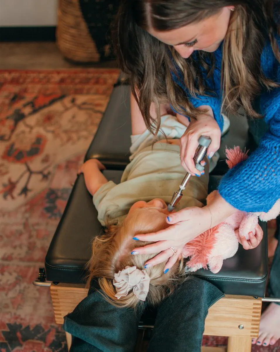 Dr. Morgan, with blue nails, gently applies a chiropractic instrument to a young patient's neck as they lie with a plush toy on the treatment table.