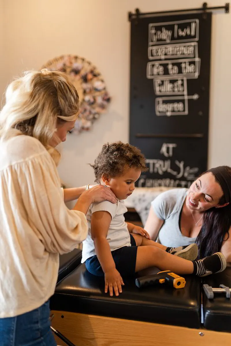 Dr. Alexis assesses a baby on a chiropractic table, while another woman smiles at the child in a comfortable, well-decorated clinic room.