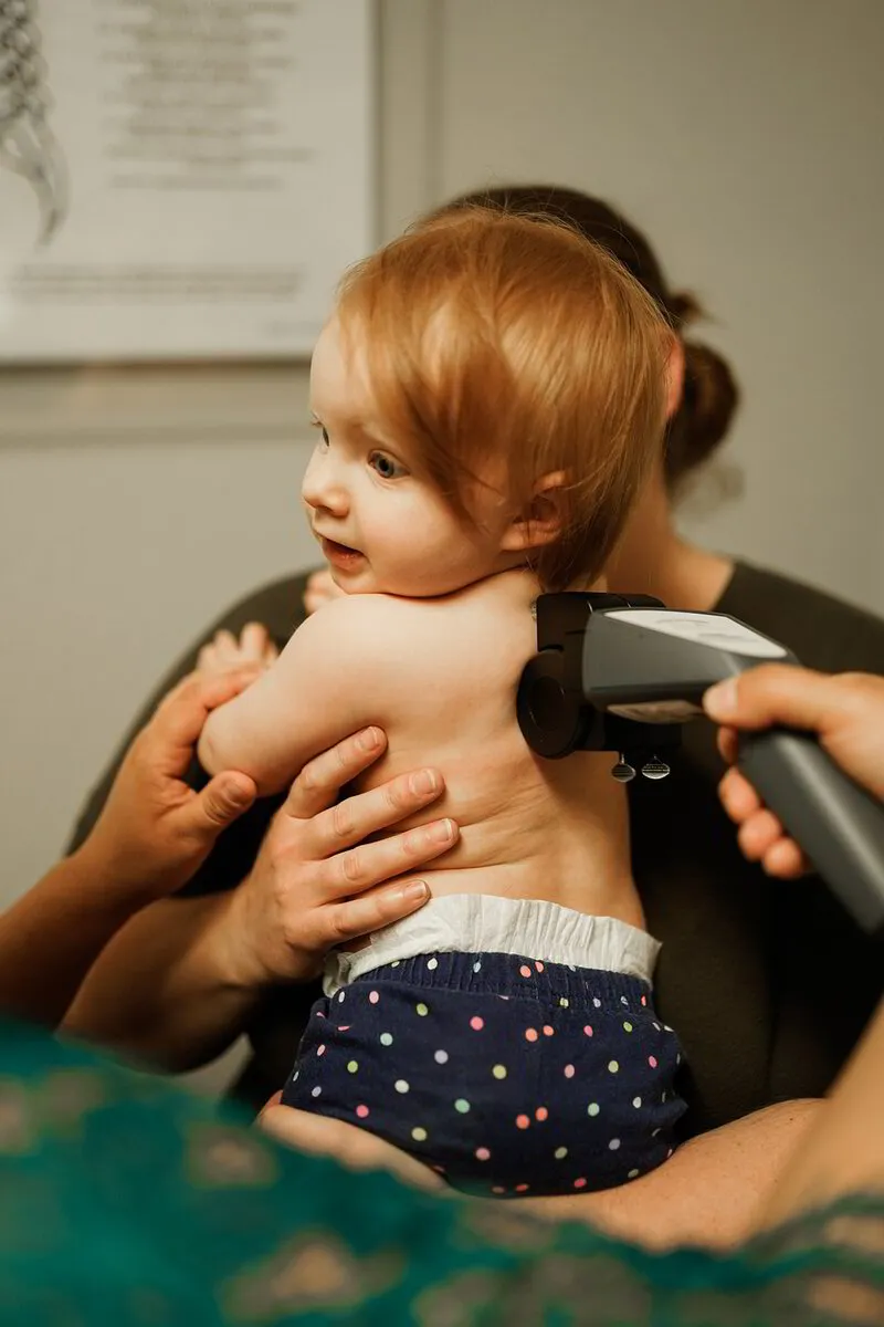 A toddler with reddish hair is held securely while receiving a gentle chiropractic treatment with a handheld device