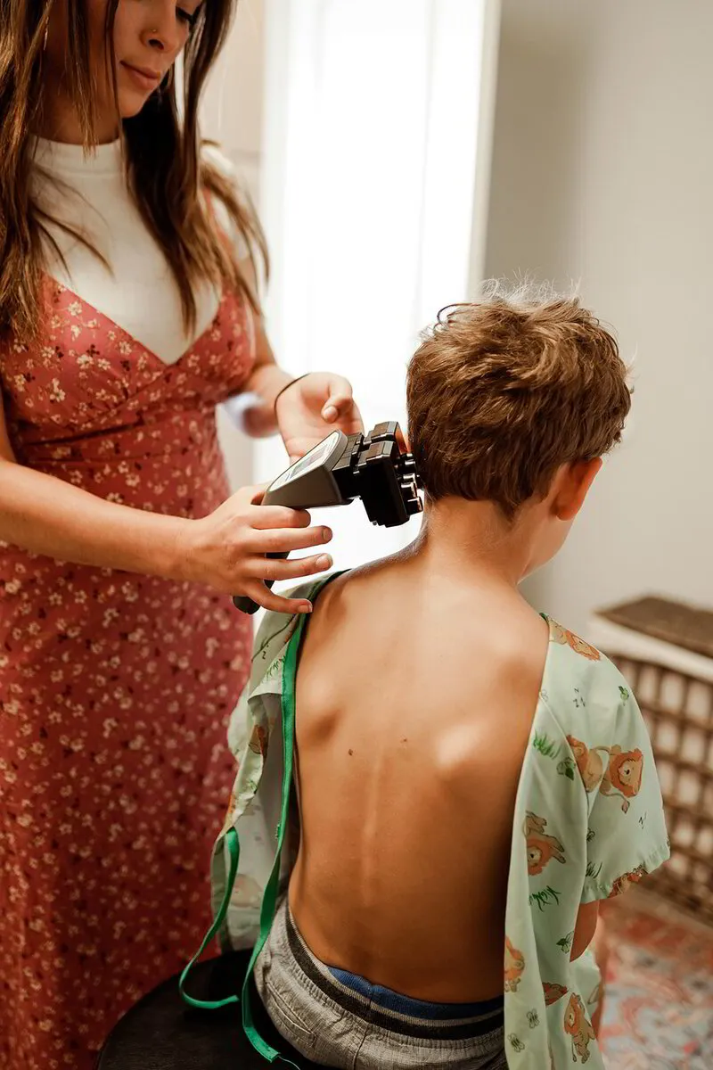 A chiropractor in a floral dress uses a spinal adjustment device on a young boy's back as he sits on a stool in a clinic with a homey ambiance.