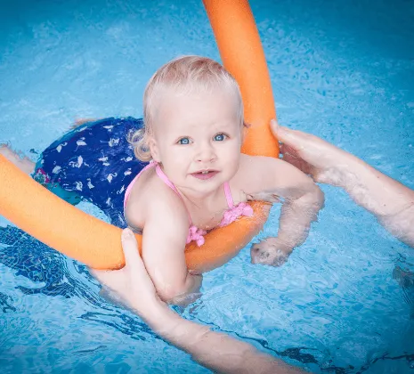 A young child swims on an orange woggle and she looks up to the camera