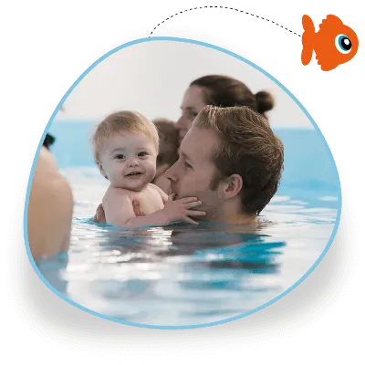 A dad holds his baby boy in the water as part of their swimming lesson, the baby is looking and smiling at the camera