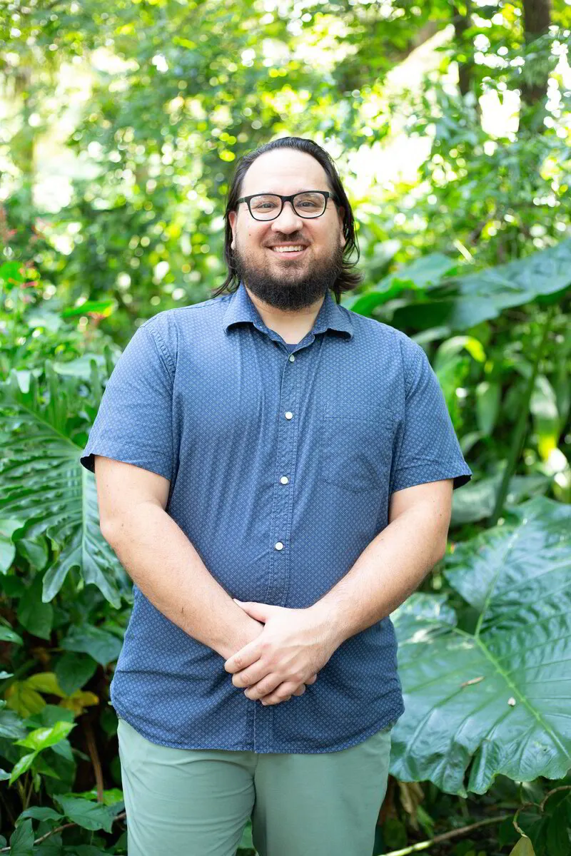 Co-Owner and Licensed Massage Therapist, Justin Mainit standing in front of foliage at Dickson Azalea Park in Orlando, Florida. 