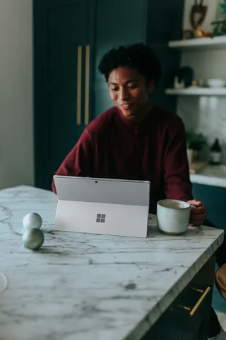 Woman in a burgundy shirt using a Surface tablet at a marble kitchen table with a cup of coffee.