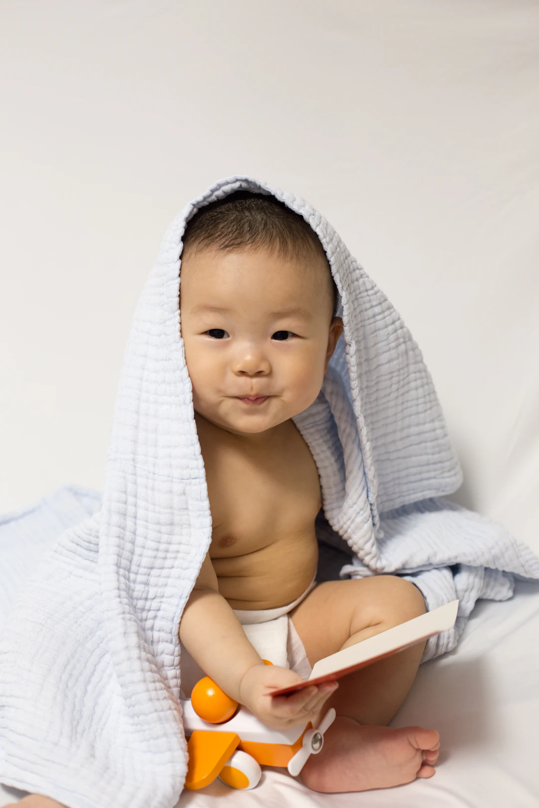 Baby wrapped in a towel holding a toy and book, sitting with a playful expression.