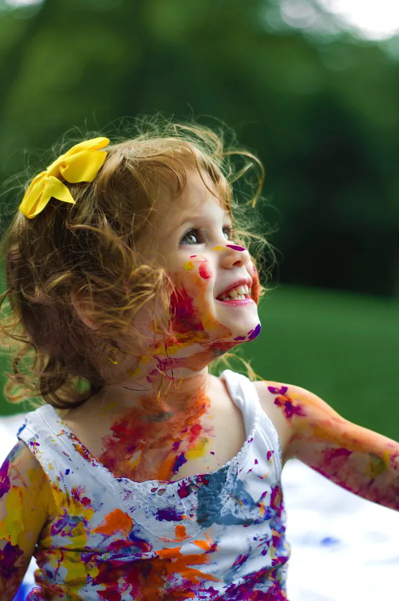 Smiling toddler with paint on face and clothes looking up outdoors.