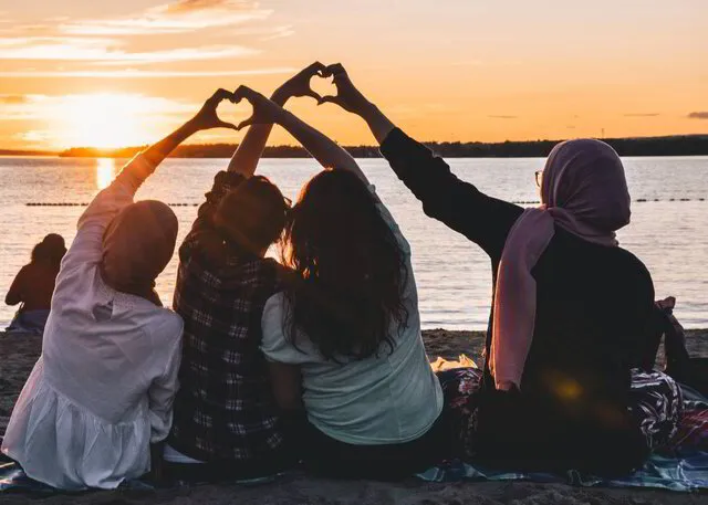 Grupp tjejer som sitter på en strand i solnedgång och gör hjärtan med händerna