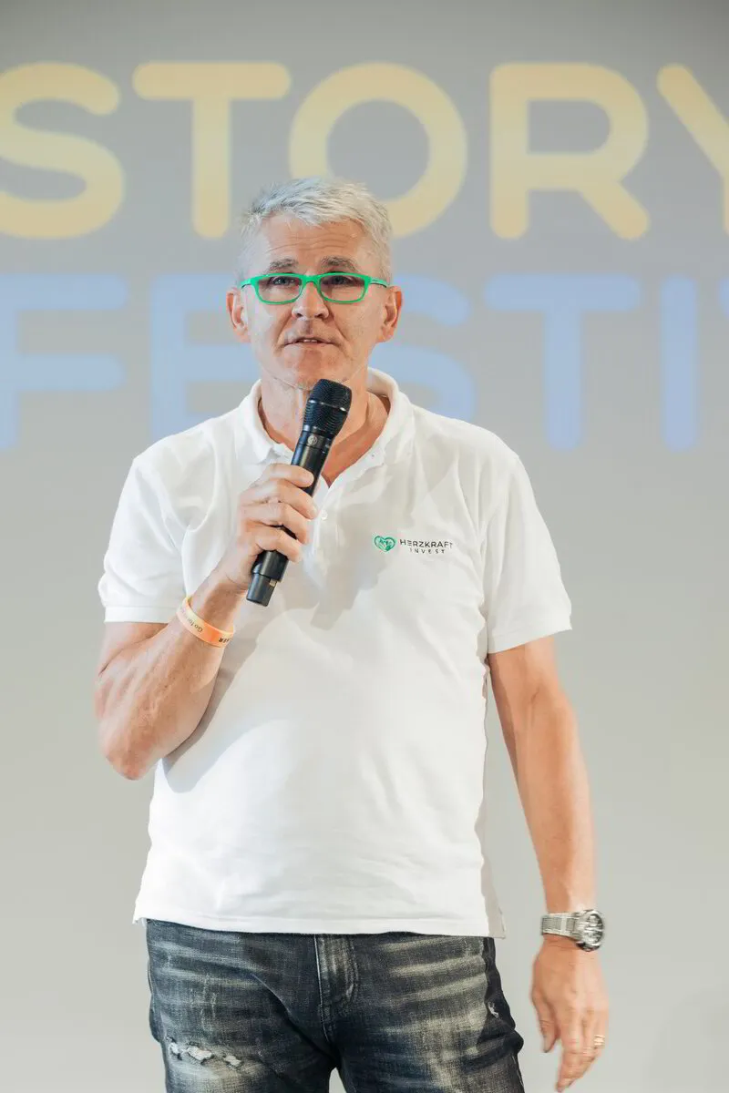 Hans Koppenhöfer mit grünen Brillengestellen, bei seiner Keynote auf dem Storyfestival von dem Institut für Storytelling (Alexander Christiani). Er trägt ein weißes Polo-Shirt mit dem Herzkraft Invest Logo und hält ein Mikrofon in seiner Hand.