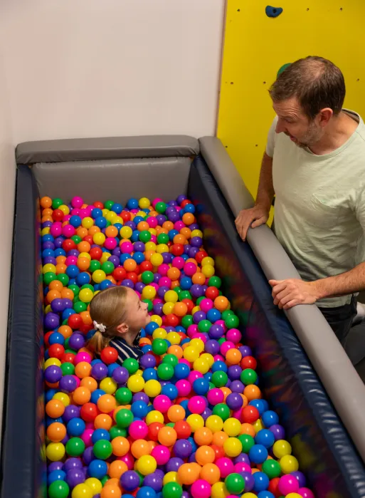 Little girl playing in a ball pit. Her physical therapist is talking with her while next to the ball pit. 